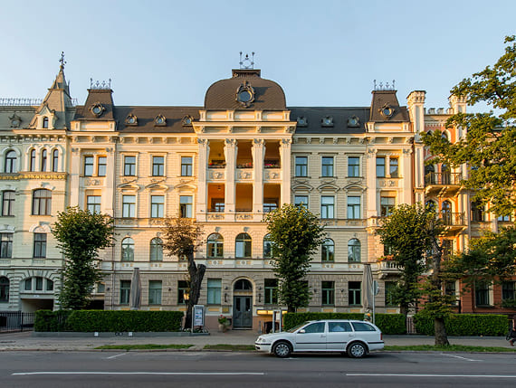 Grand historic building with ornate facade and central columns, facing a tree-lined street with a parked car in front.